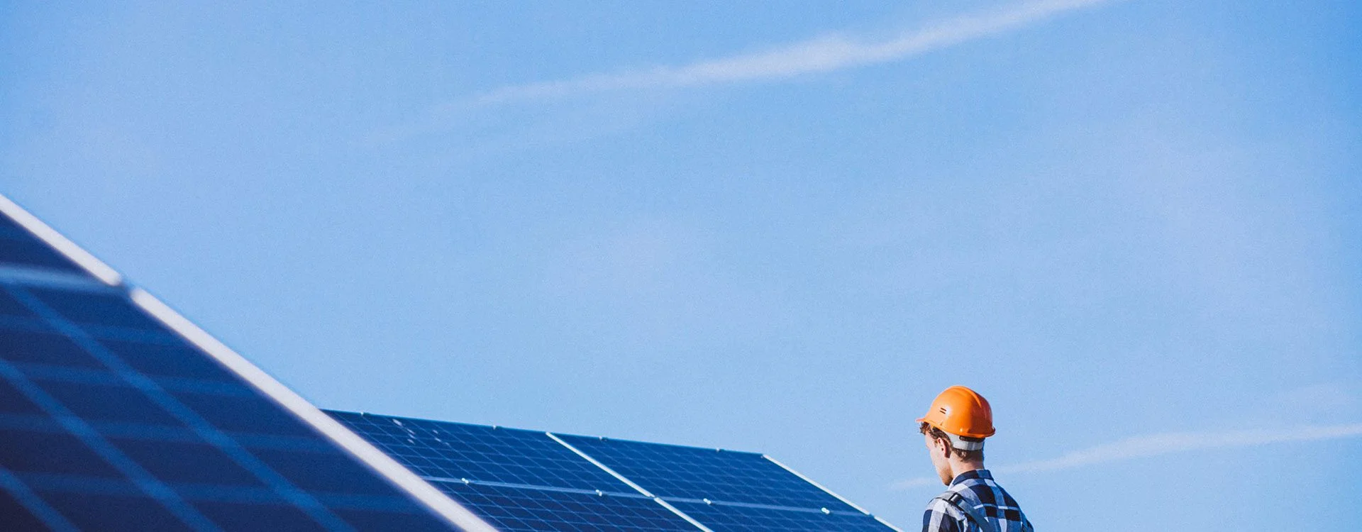 Worker in hard hat inspecting solar panels under a blue sky.