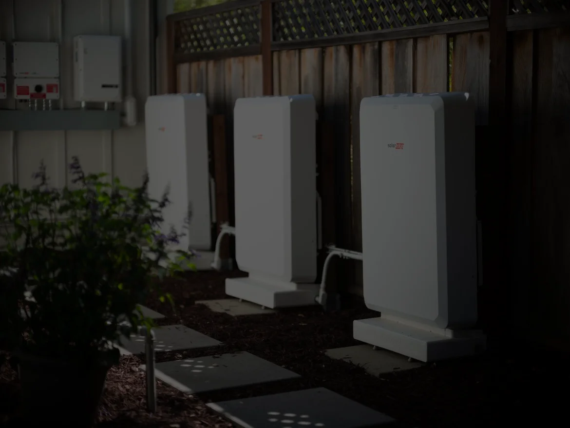 Three white SolarEdge battery storage units installed outdoors next to a wooden fence.